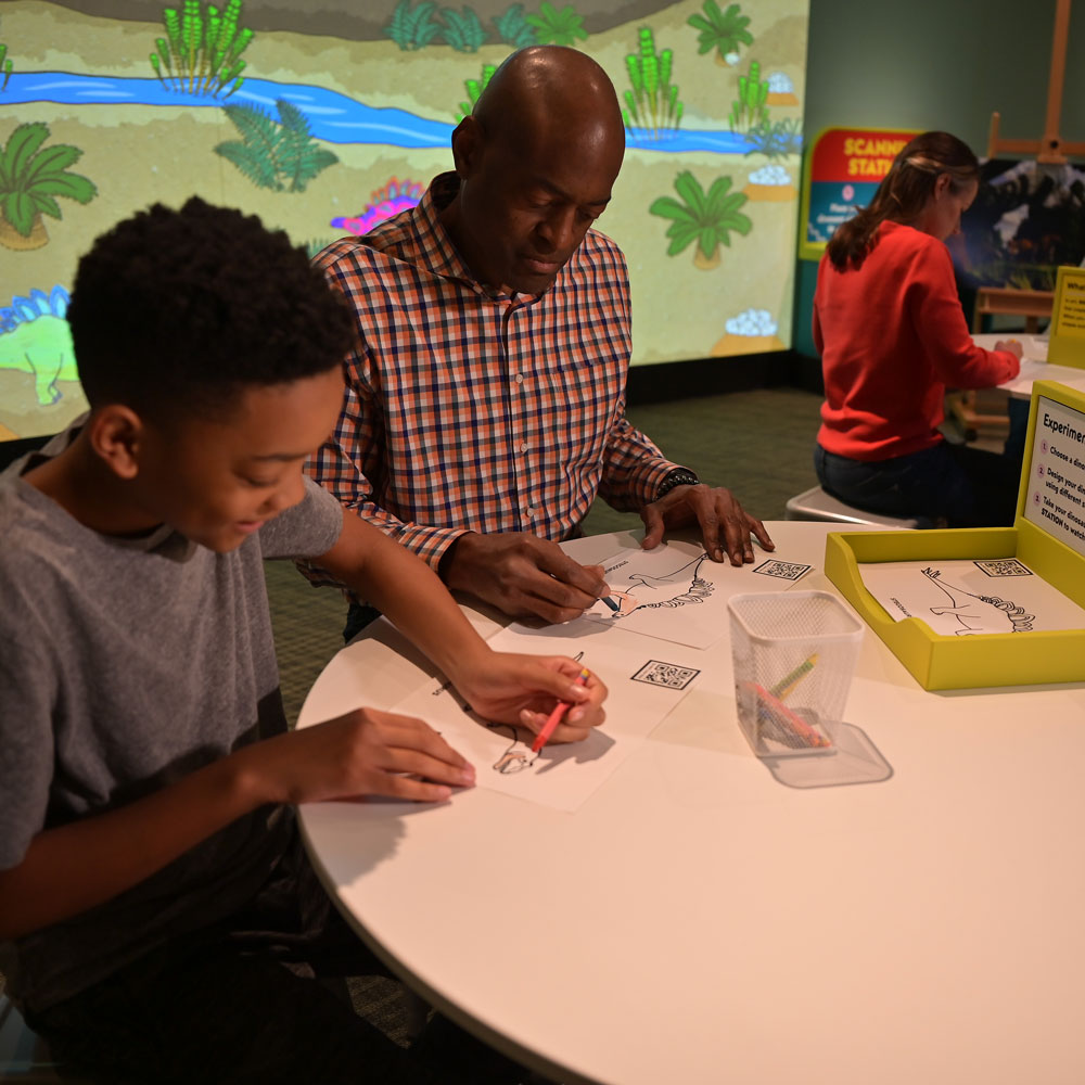 A grown-up and a young man sitting at a table coloring dinosaur pictures. In the background is a projection screen with other visitors' dinosaur designs on display.